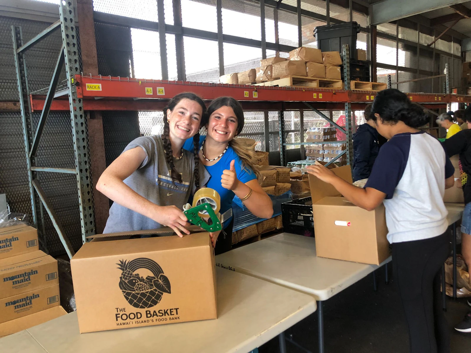 Two teen travelers give a thumbs-up while taping a Food Basket box shut in a warehouse lined with shelves