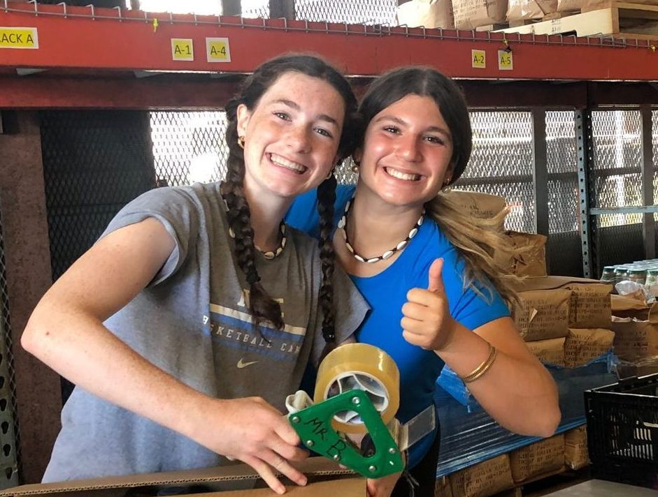 Students smile and give a thumbs-up while packing tape at a food pantry warehouse