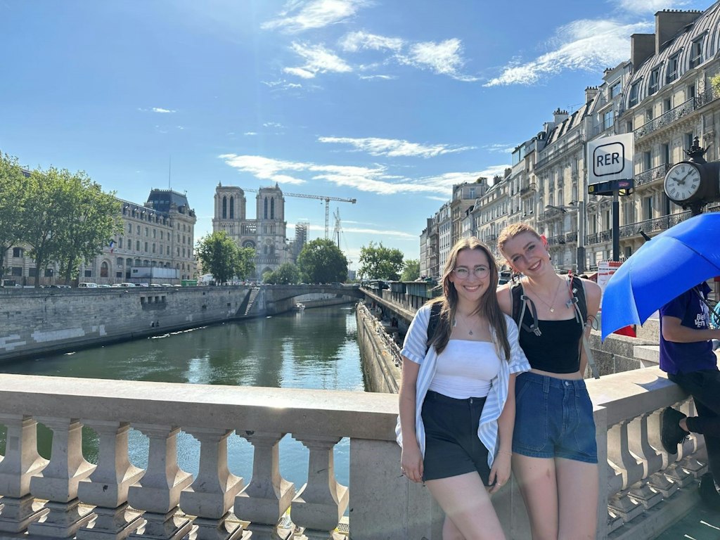 Two smiling friends pose on a bridge in Paris with Notre-Dame Cathedral and the Seine River in the background.