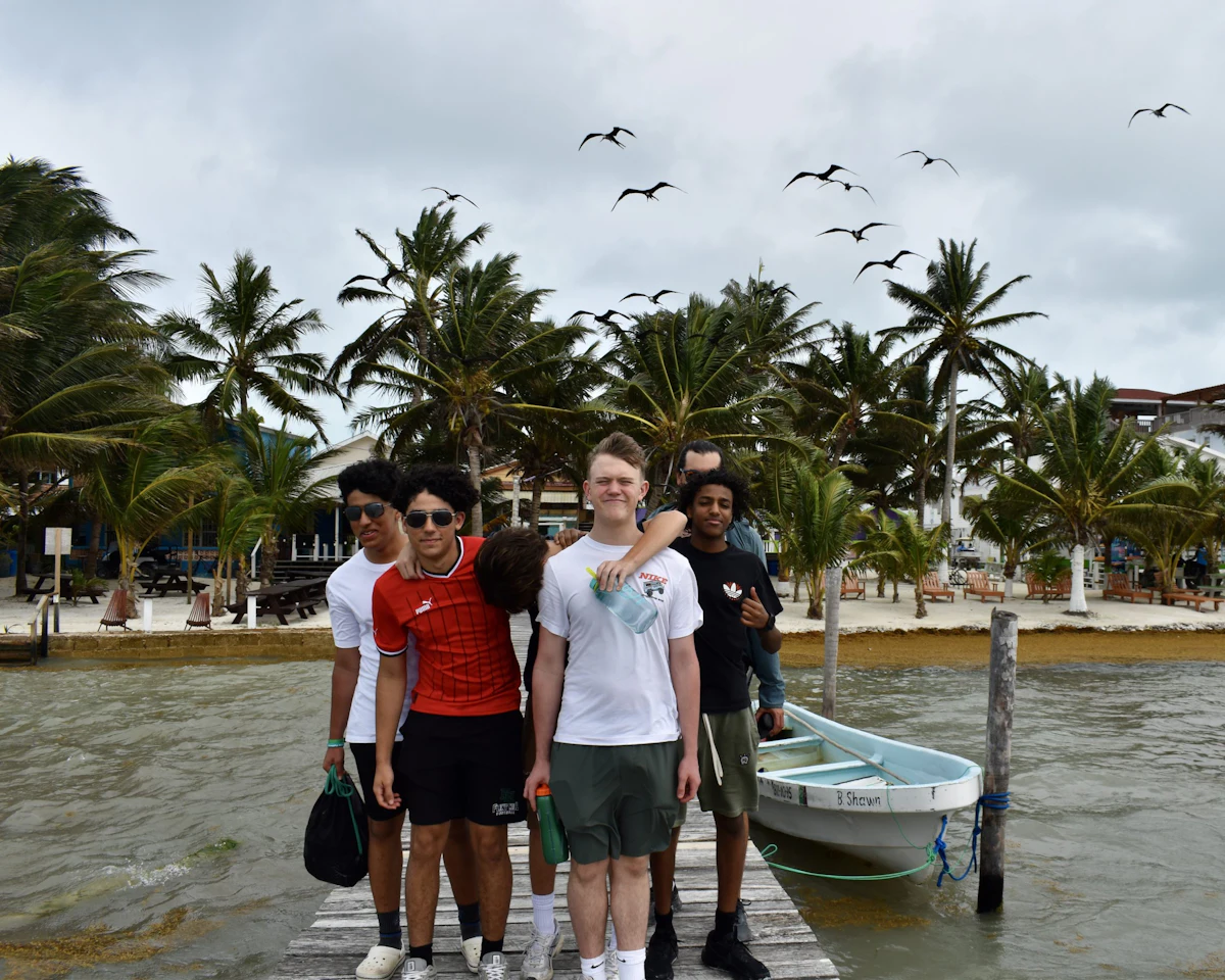Five teen boys on a Rustic Pathways program stand on a wooden dock in Belize, with palm trees and frigatebirds behind them