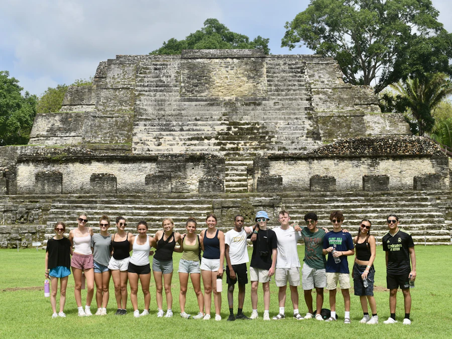 A school group stands together on the grass in front of a stepped Maya temple in Belize