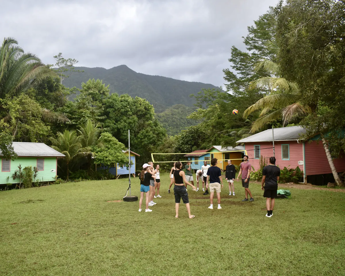 Students playing volleyball on a grass court between painted cabins, forested hills behind them