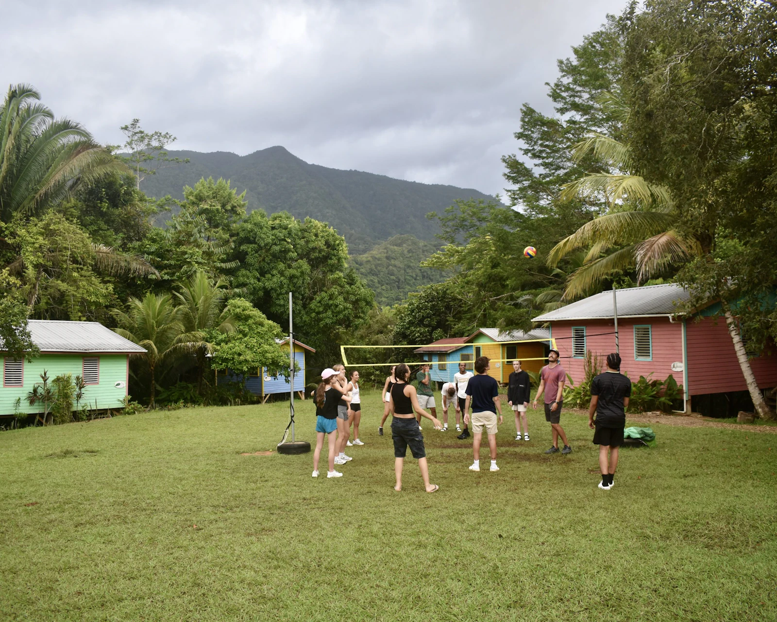Students play volleyball on the grass beside colorful cabins in the Belize rainforest, with mountains behind