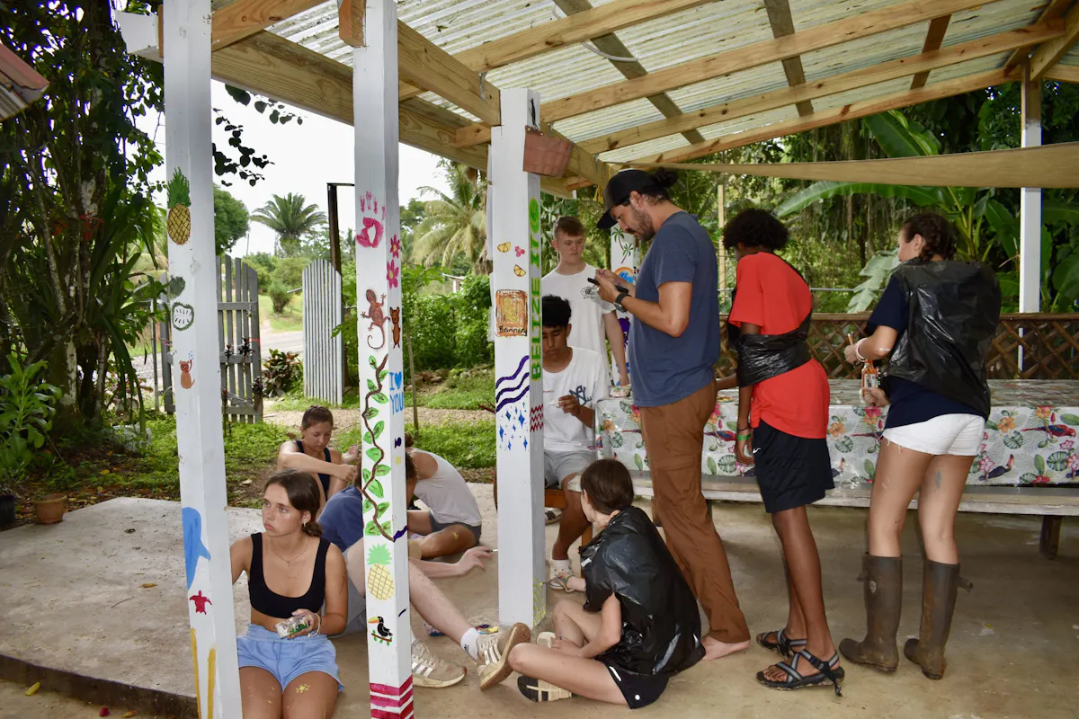 Students painting patterns onto the posts of an open-sided shelter, a Rustic Pathways staff member working among them
