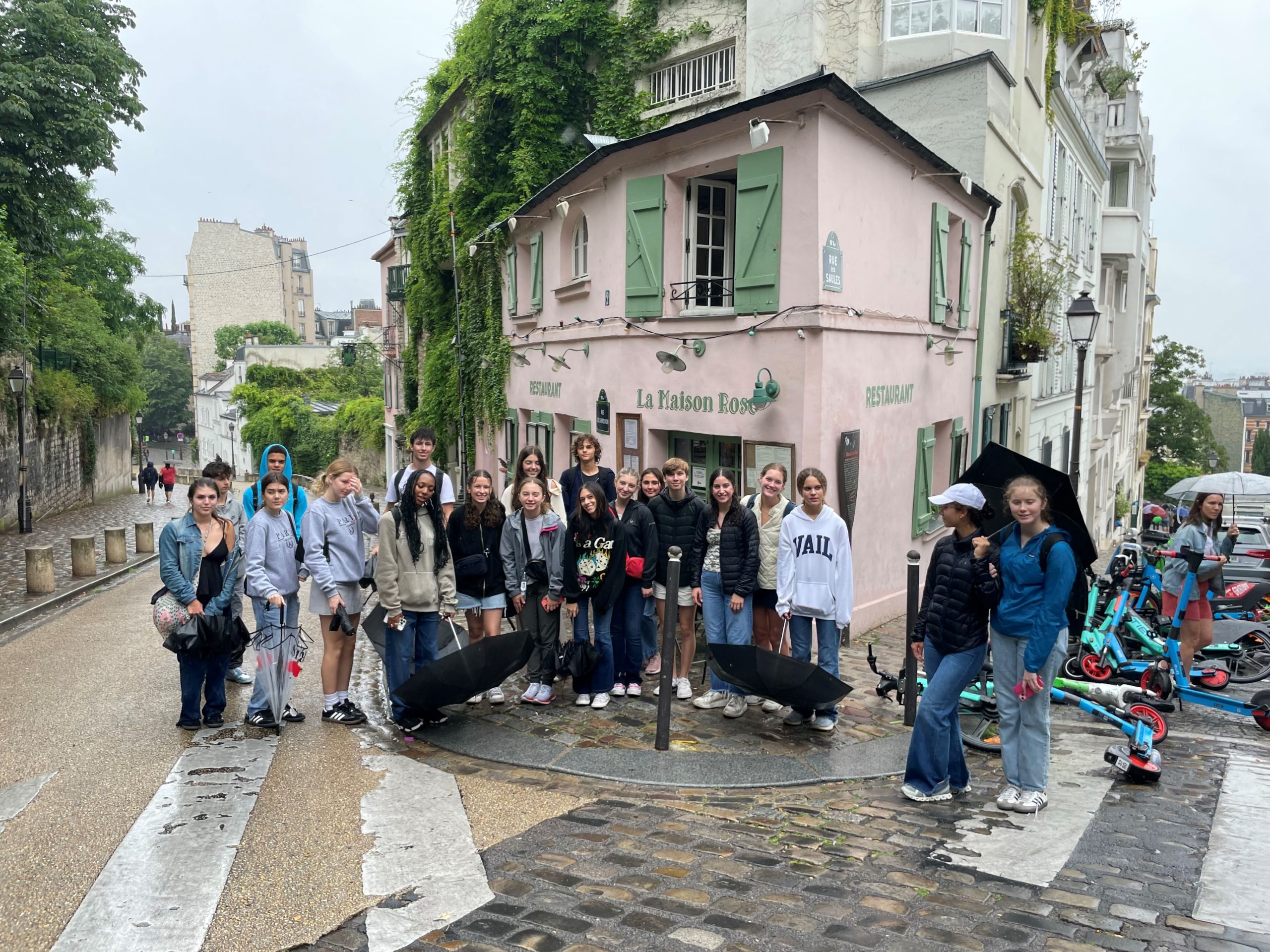 A group of students gathers outside a pink corner cafe on a cobblestone Montmartre street, framed by ivy-covered buildings.