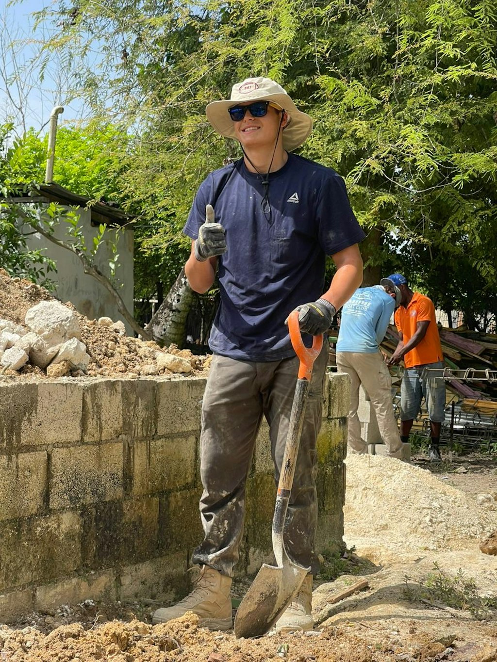 A working student smiling in to the camera with thumbs up. The setting shows a construction site, that the students are working on.