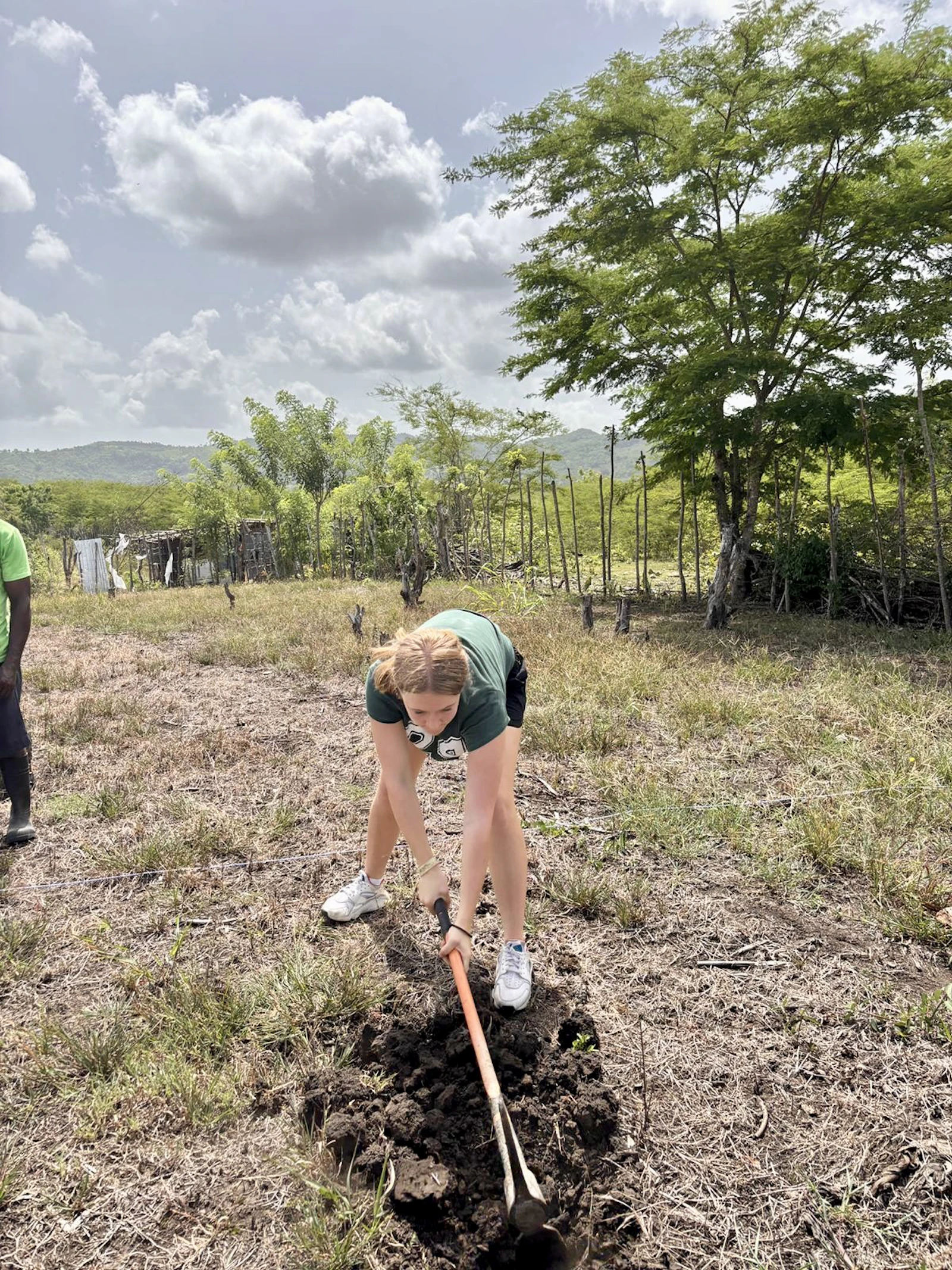 A teen traveler digs a planting hole with a long iron bar in an open field in the Dominican Republic
