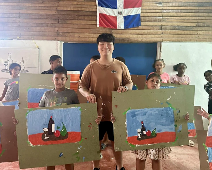 Students hold up paintings in a classroom with a Dominican Republic flag on the wall