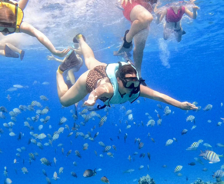 A teen traveler in a turquoise snorkel vest swims down through a school of striped reef fish in the Dominican Republic