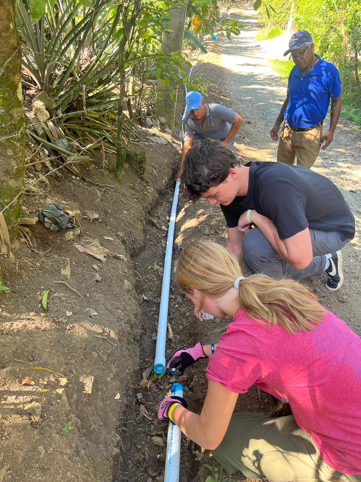 Students kneel along a dirt trail installing a water pipe as community members walk by