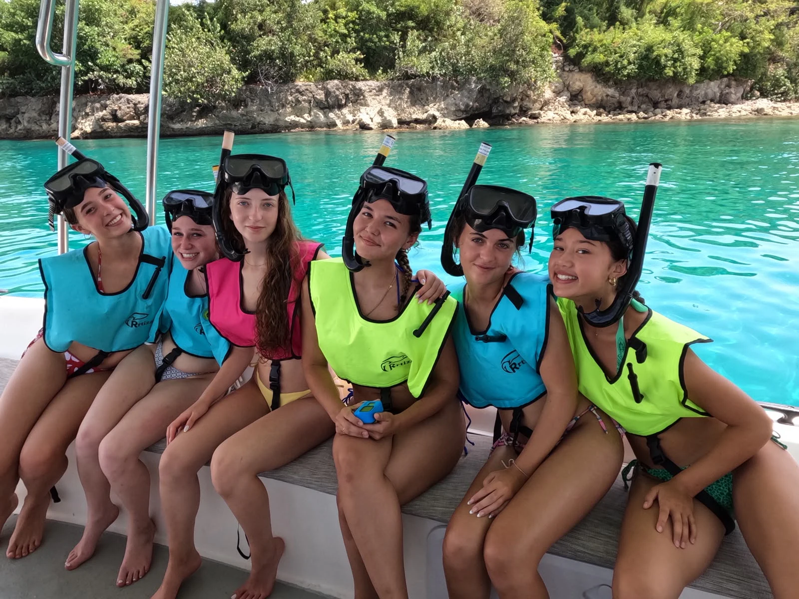 Seven teen travelers in snorkel vests and masks sit along the side of a boat above turquoise water in the Dominican Republic