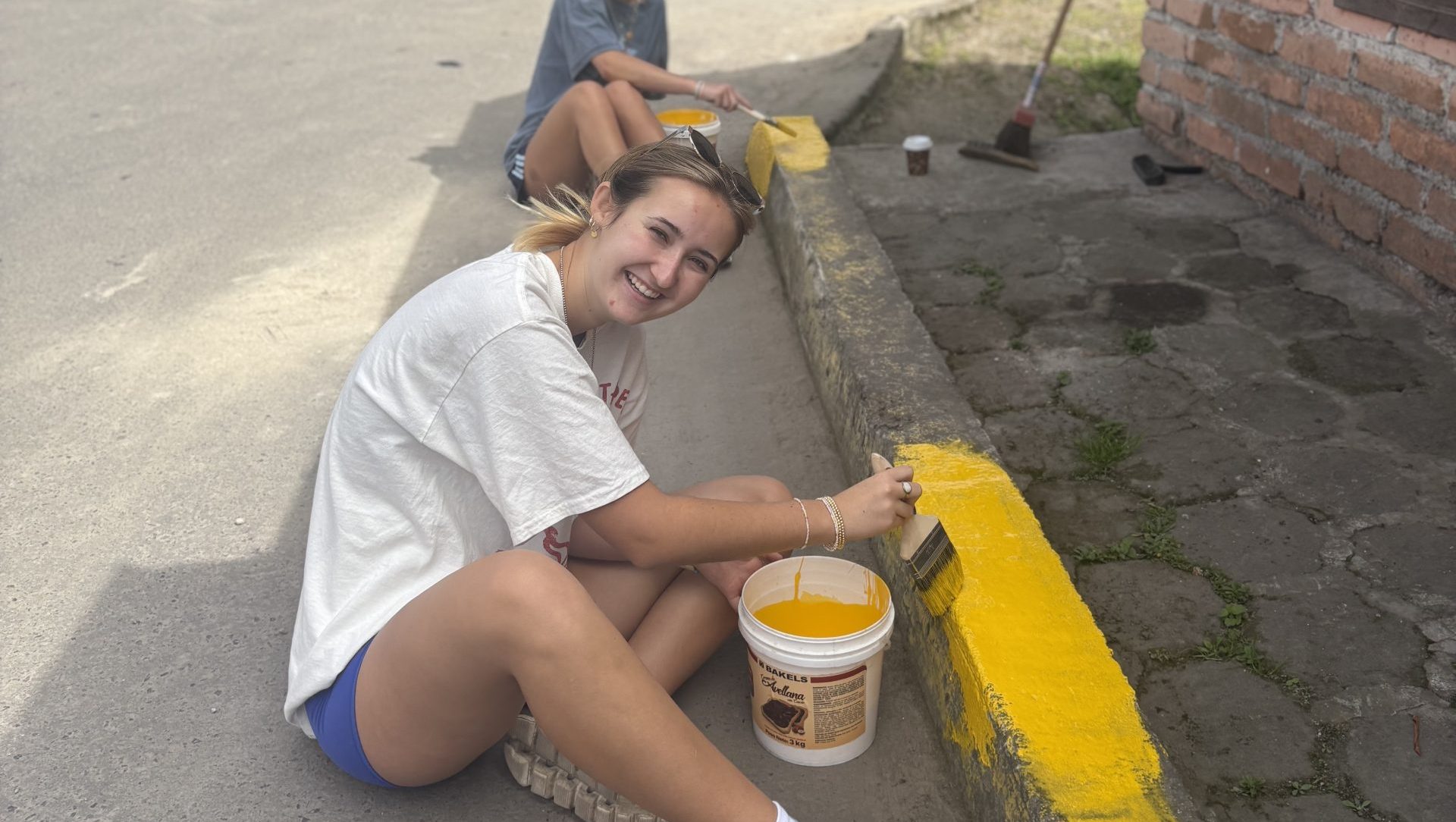 A student smiles while painting a curbstone yellow with a bucket of paint
