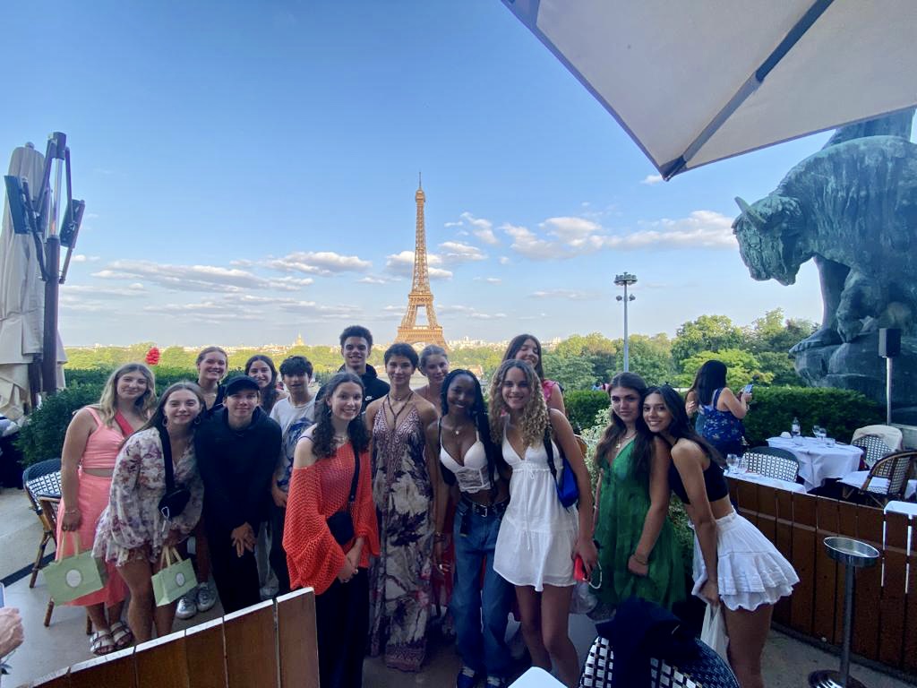 A group of students smiles on a rooftop terrace with the Eiffel Tower rising in the distance behind them.