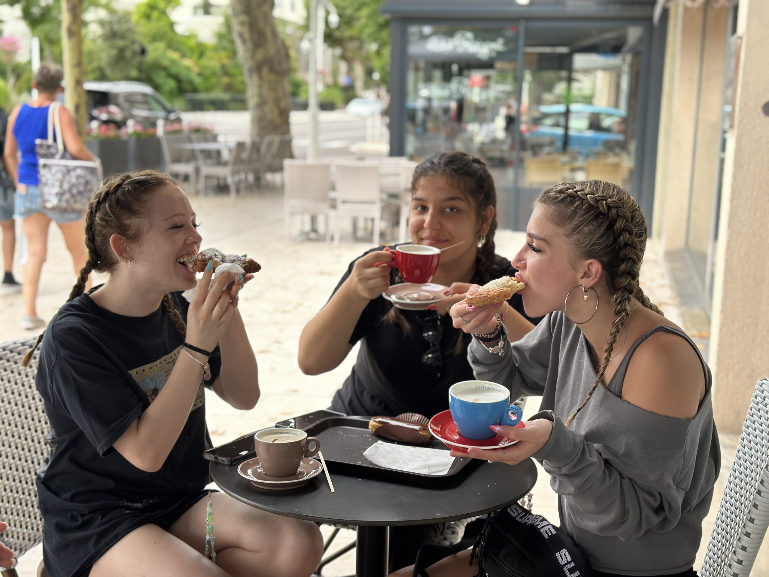 Three students laugh and share pastries and coffee at an outdoor café table in France.