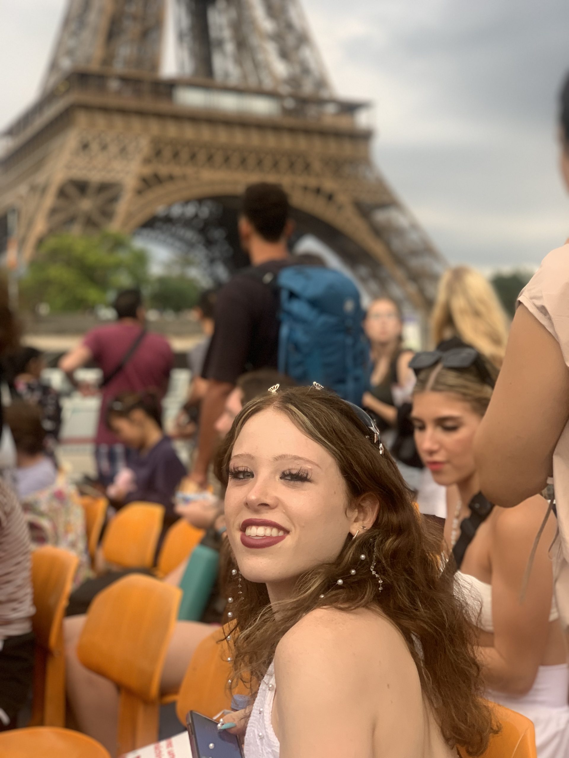 A student glances back at the camera with the Eiffel Tower's iron latticework and a crowd of visitors behind her.