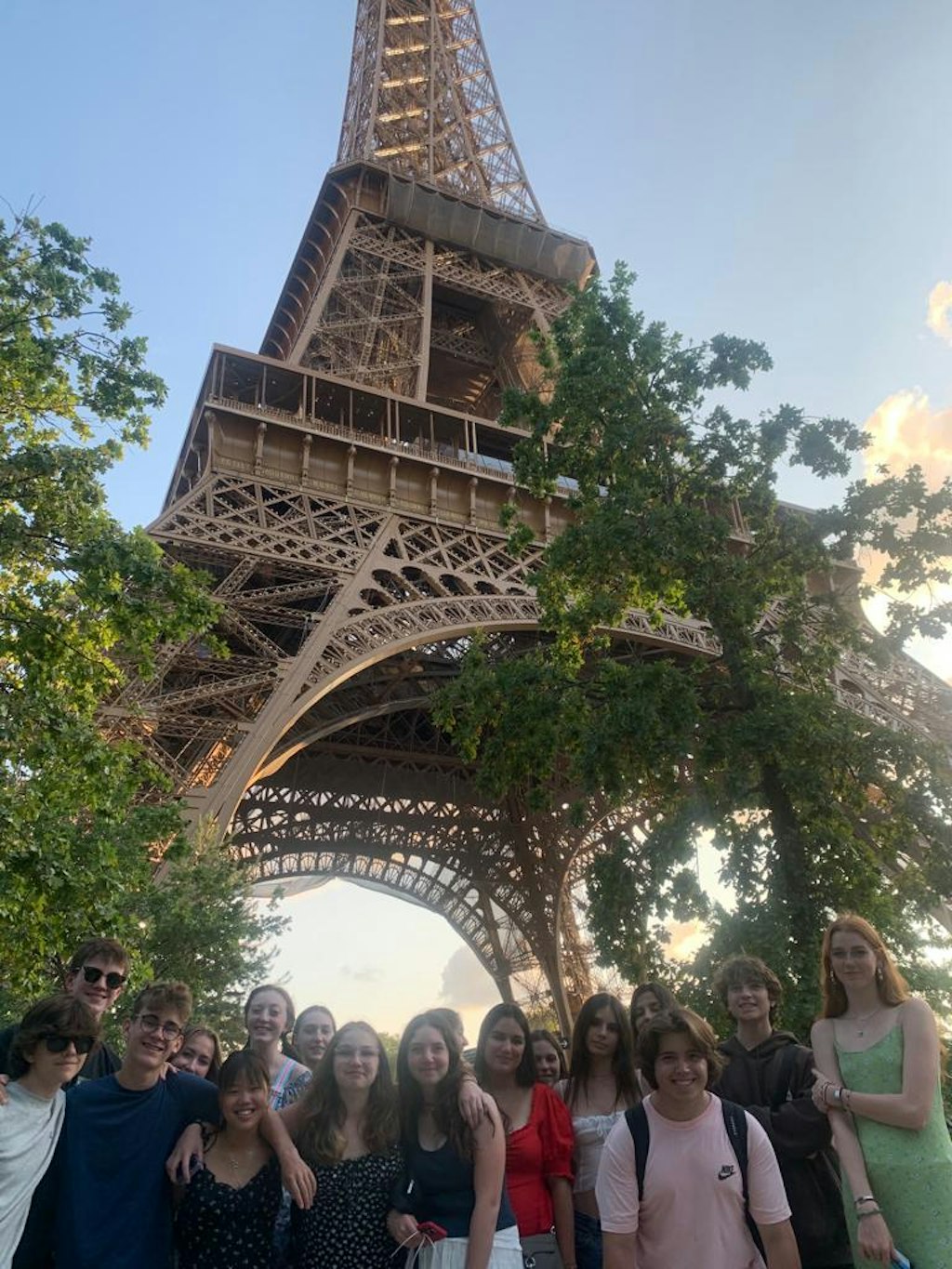 A group of students stands in front of the Eiffel Tower, smiling for the camera. The iconic tower looms in the background, its iron lattice structure visible against a clear blue sky.