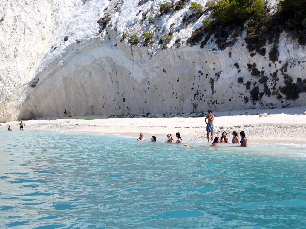 A group of people enjoying the crystal-clear waters and sandy beach along the rocky coastline of a secluded spot in Greece. White cliffs rise steeply behind them, with sparse greenery adding contrast to the pristine scene. Fun Facts About Greece