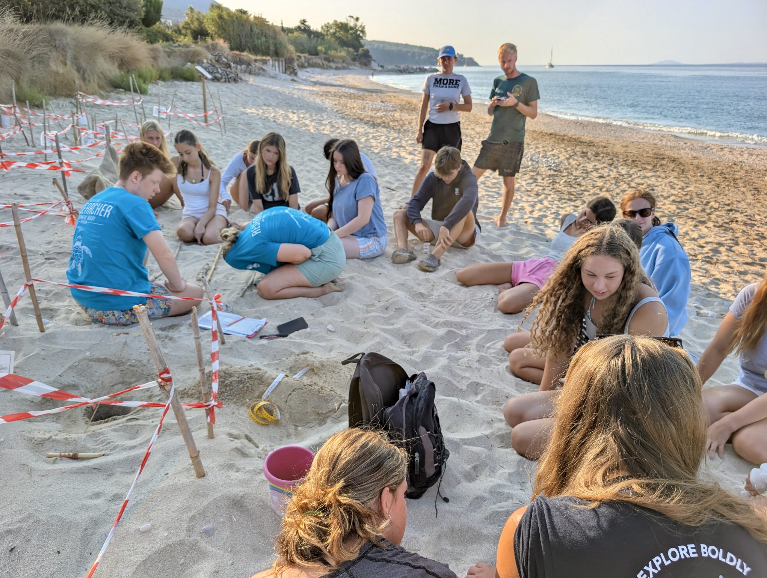 Students sit in a circle on a sandy beach around a taped-off nesting site during a turtle conservation briefing