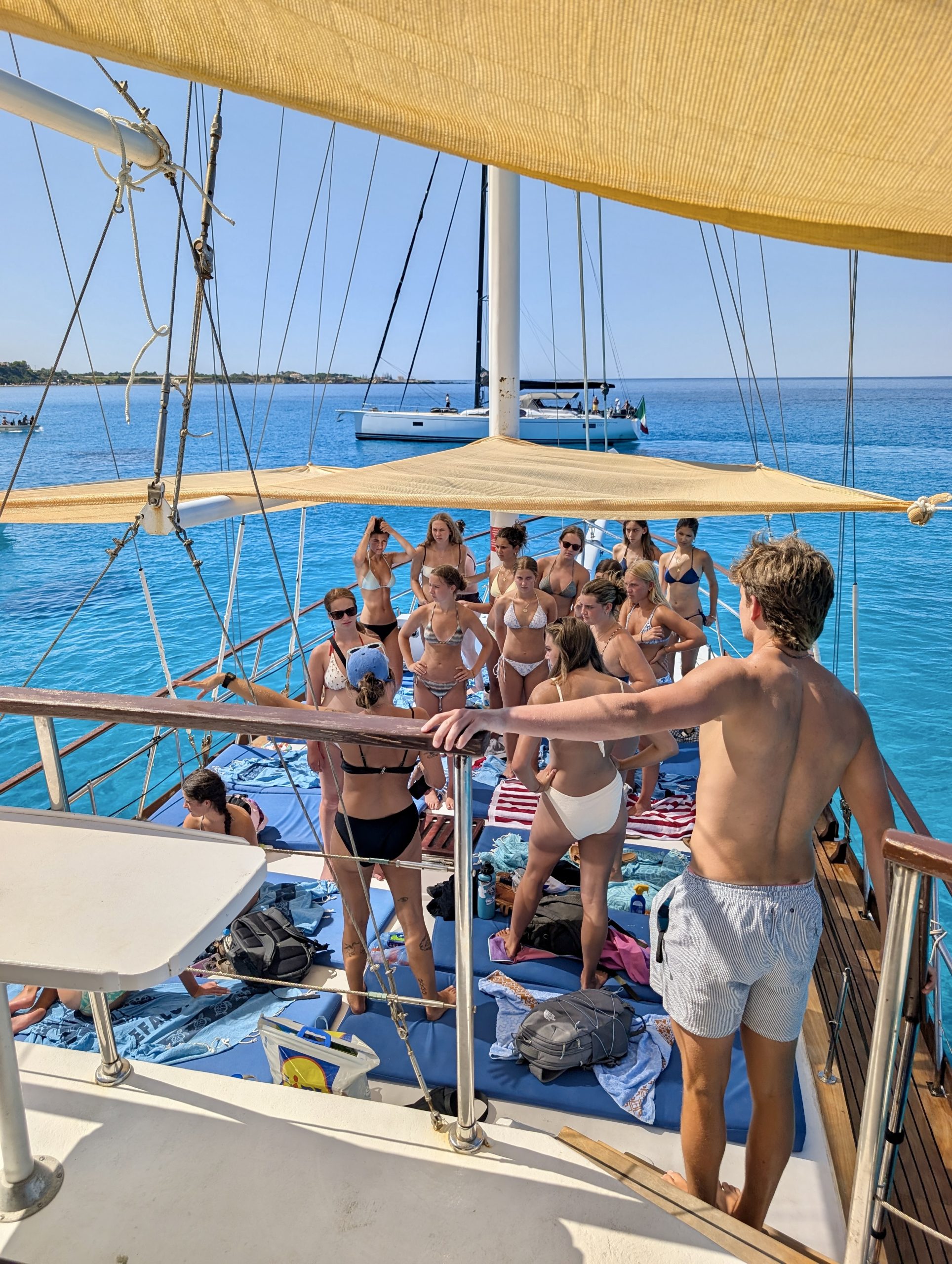 Students gather on a sailboat deck under a shade canopy over clear blue water