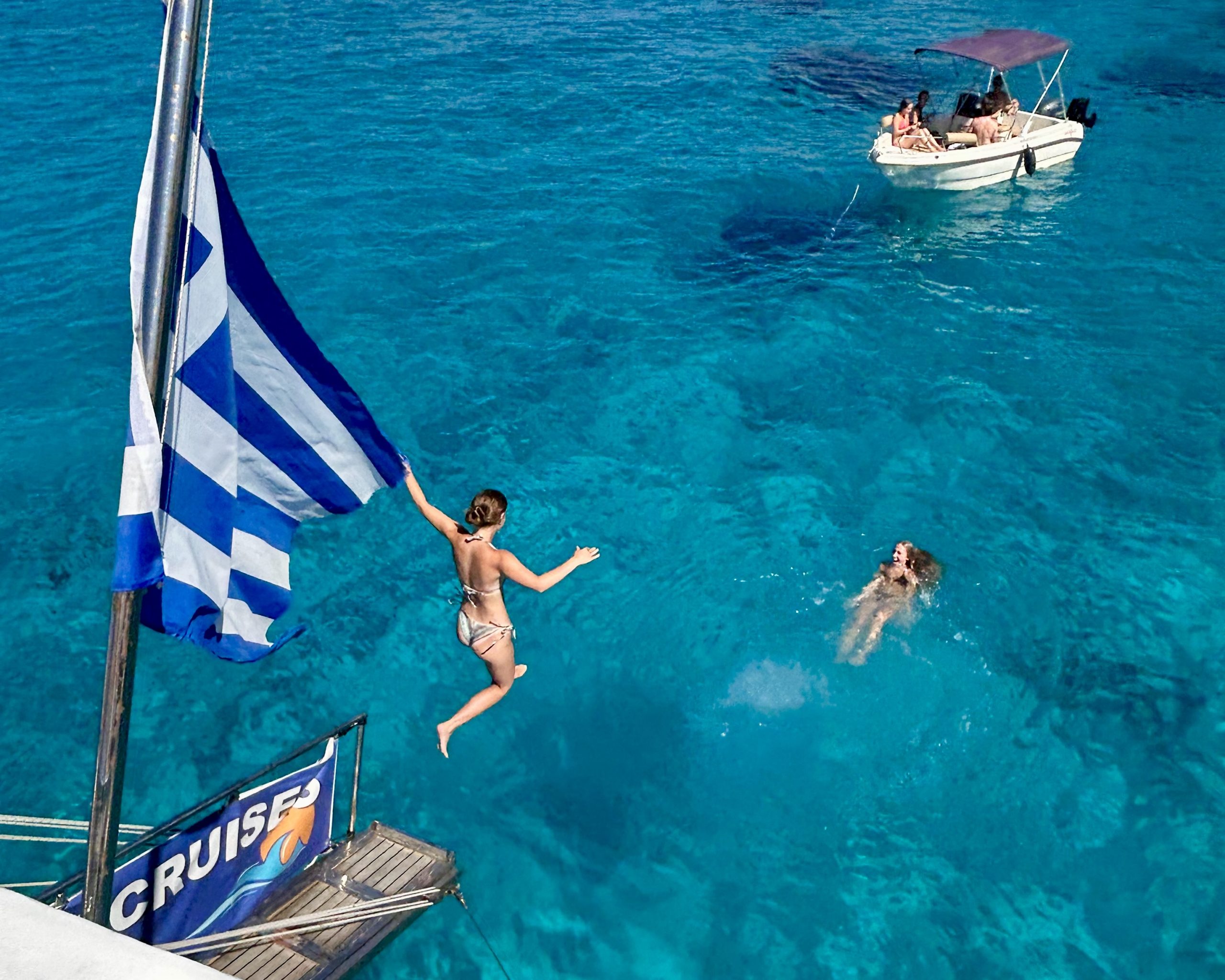 A girl jumps from a boat into bright turquoise water in Greece, holding a Greek flag, while friends watch nearby from another boat.