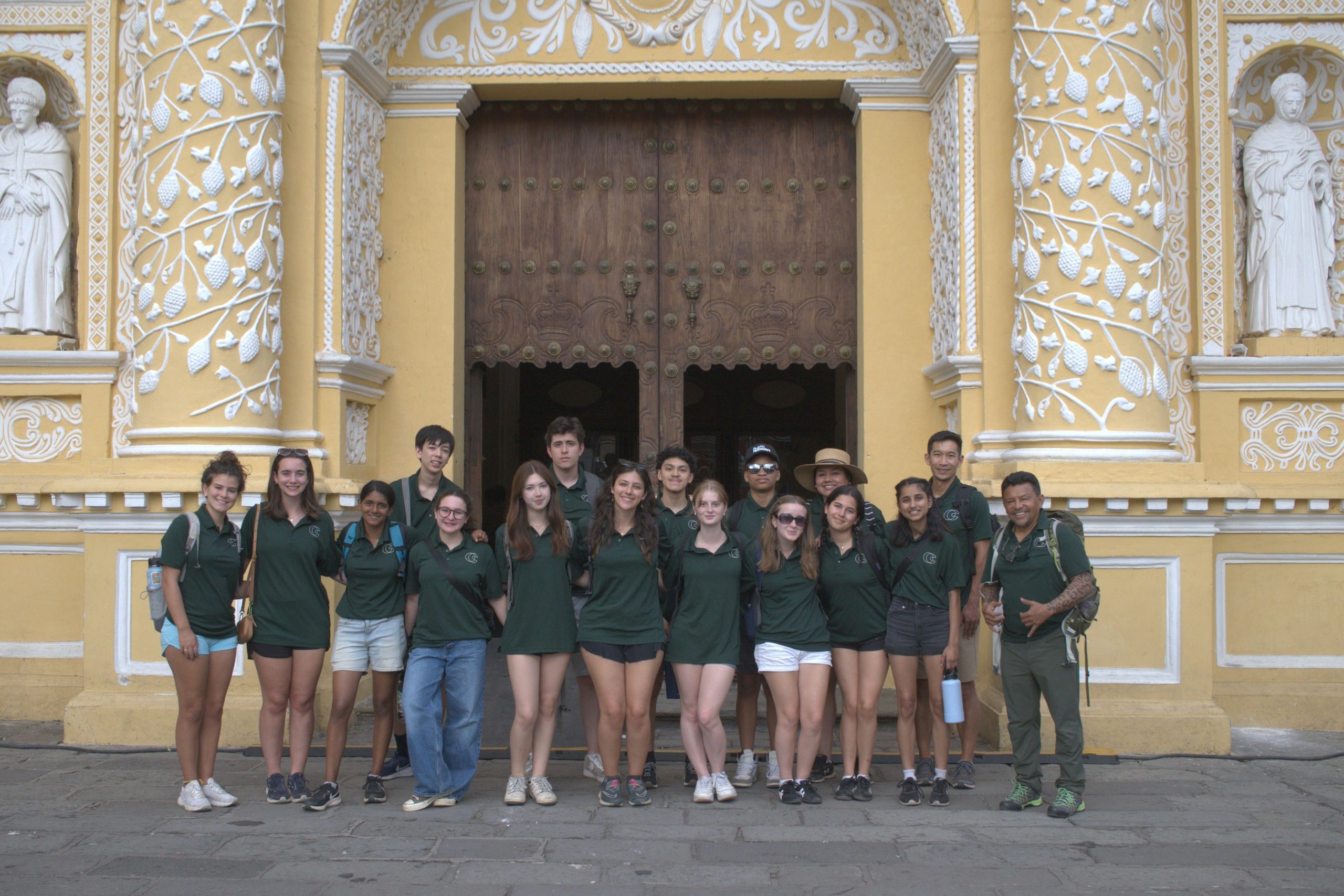 A school group in matching green polo shirts standing with two leaders in front of a yellow baroque church facade worked in white plaster.