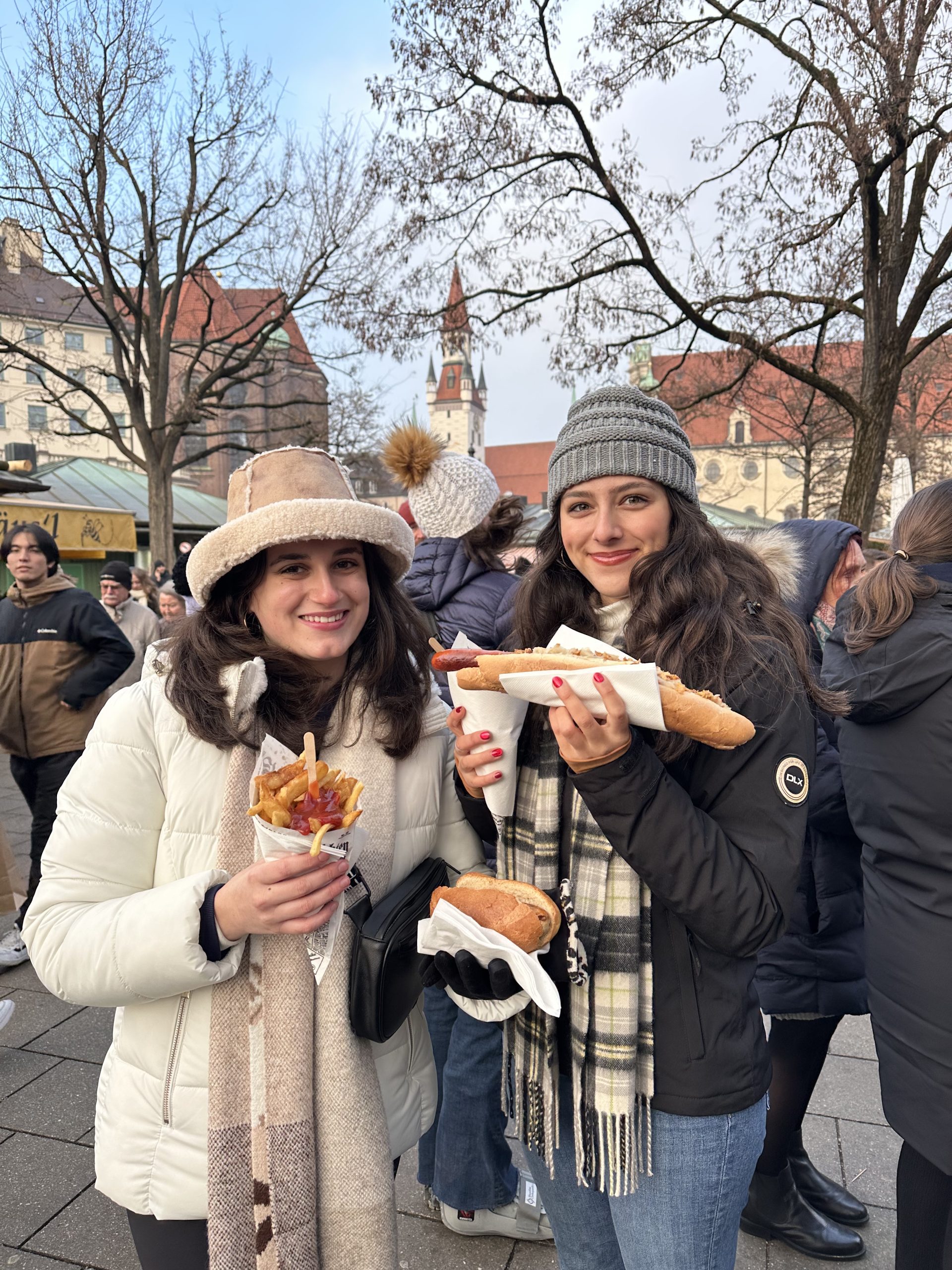 Two teen travelers enjoying local German street food at an outdoor market in Munich, smiling and holding bratwurst and fries, part of a student travel to Germany winter program.