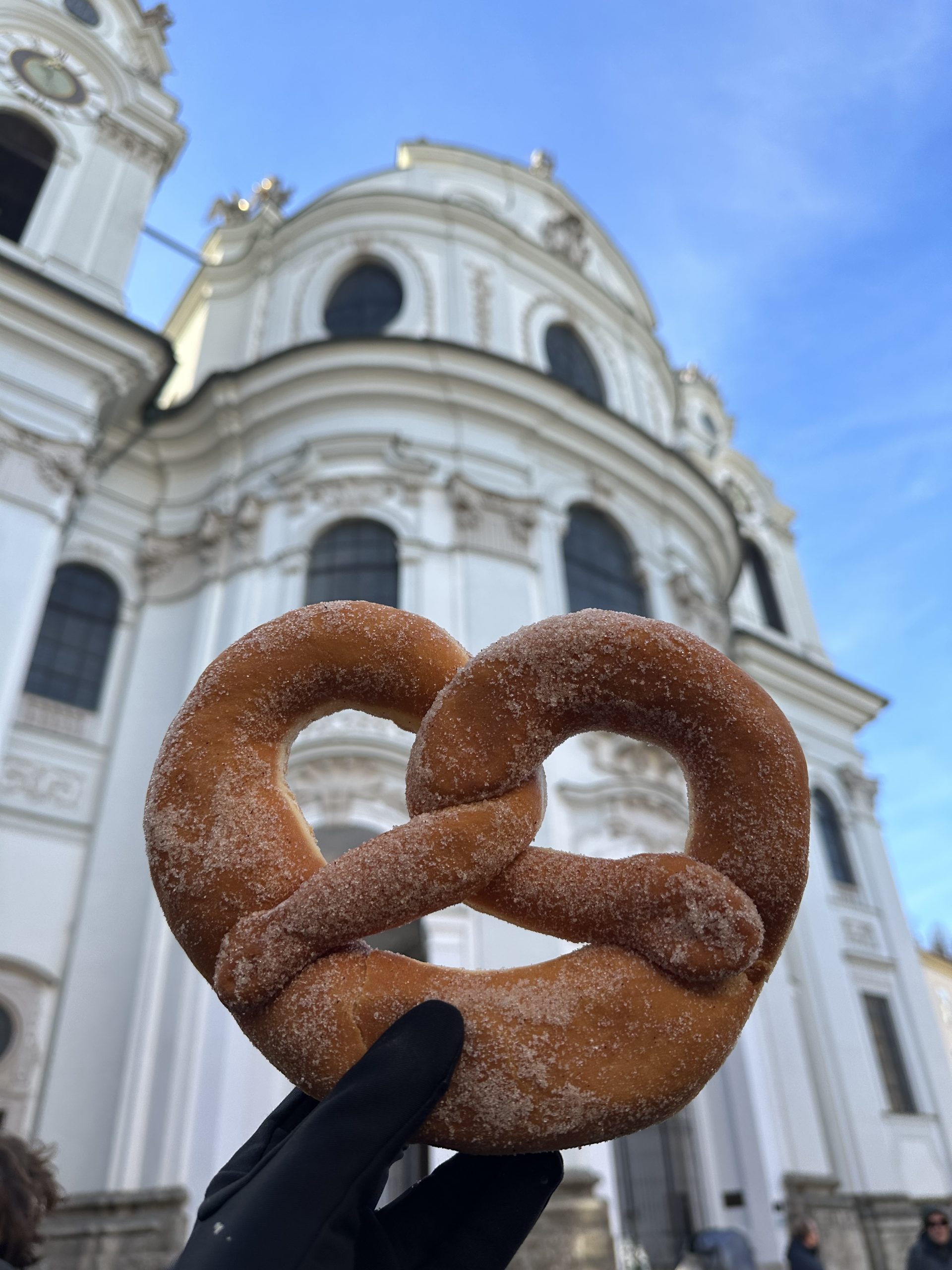 A sugar-dusted pretzel held up in front of a historic baroque church in Germany, capturing the local flavors experienced by student travelers on a teen travel adventure.