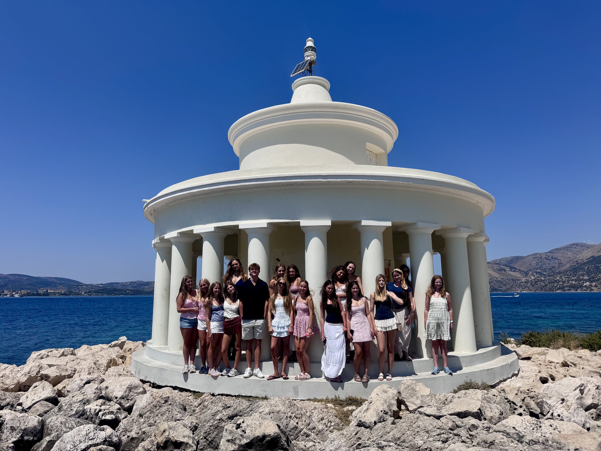 A school group stands inside the white circular colonnade of the Saint Theodore lighthouse at Argostoli on Kefalonia, with the sea behind.