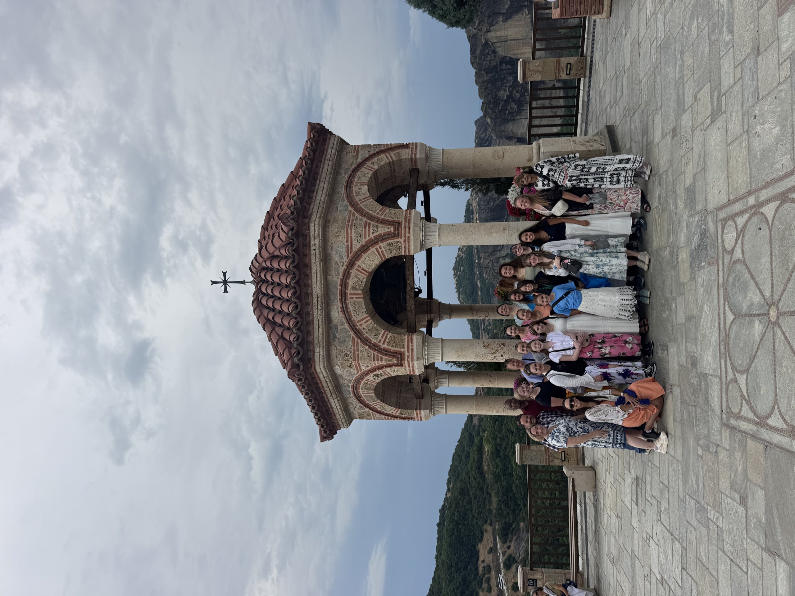 A school group gathered under the tiled dome of a monastery bell tower at Meteora, with the rock pinnacles behind.