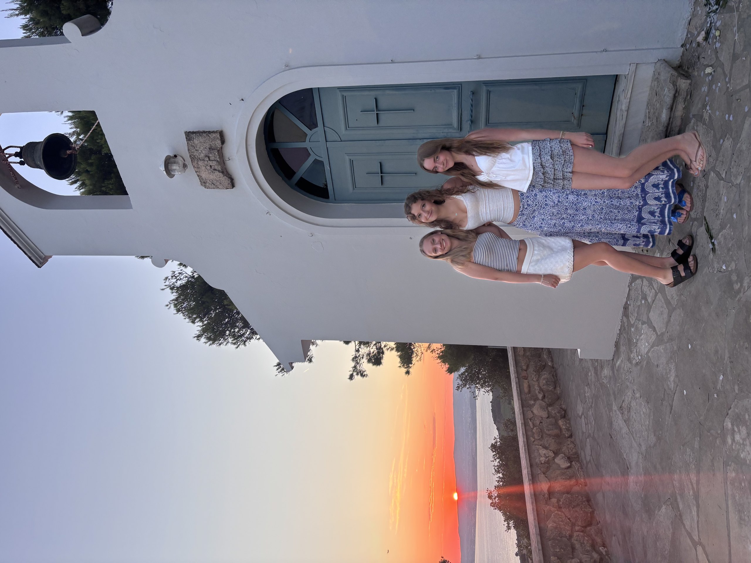 Three students stand beside the blue door of a whitewashed Greek chapel as the sun sets over the sea behind them.