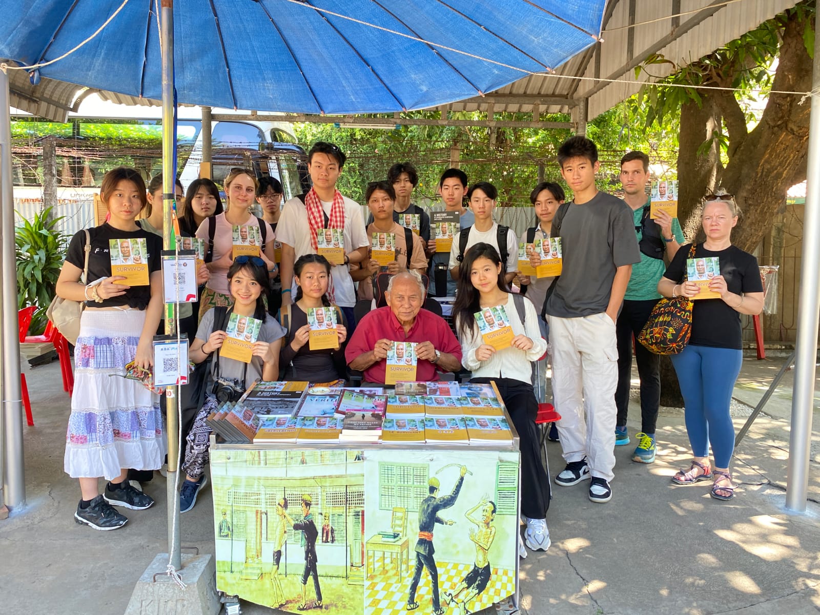 Students gather under a blue umbrella holding illustrated cards with a local artist seated at a painted stand