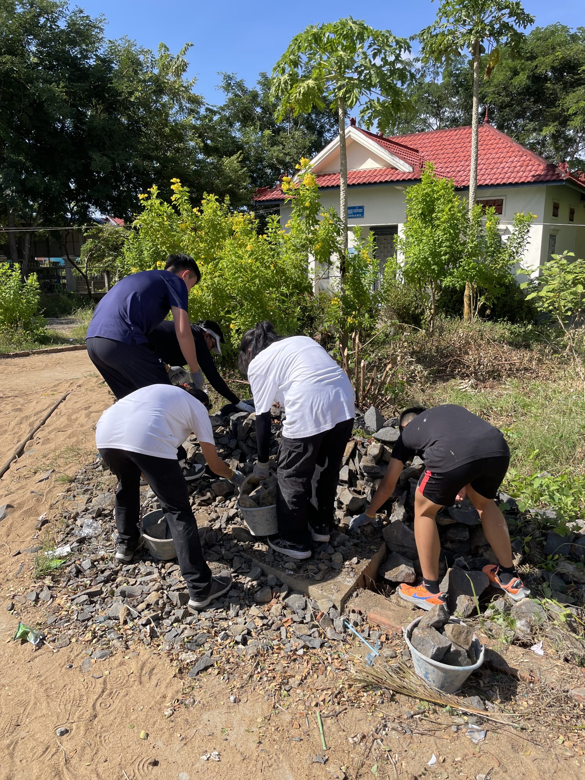 Students gather stones into buckets along a dirt path near a village house