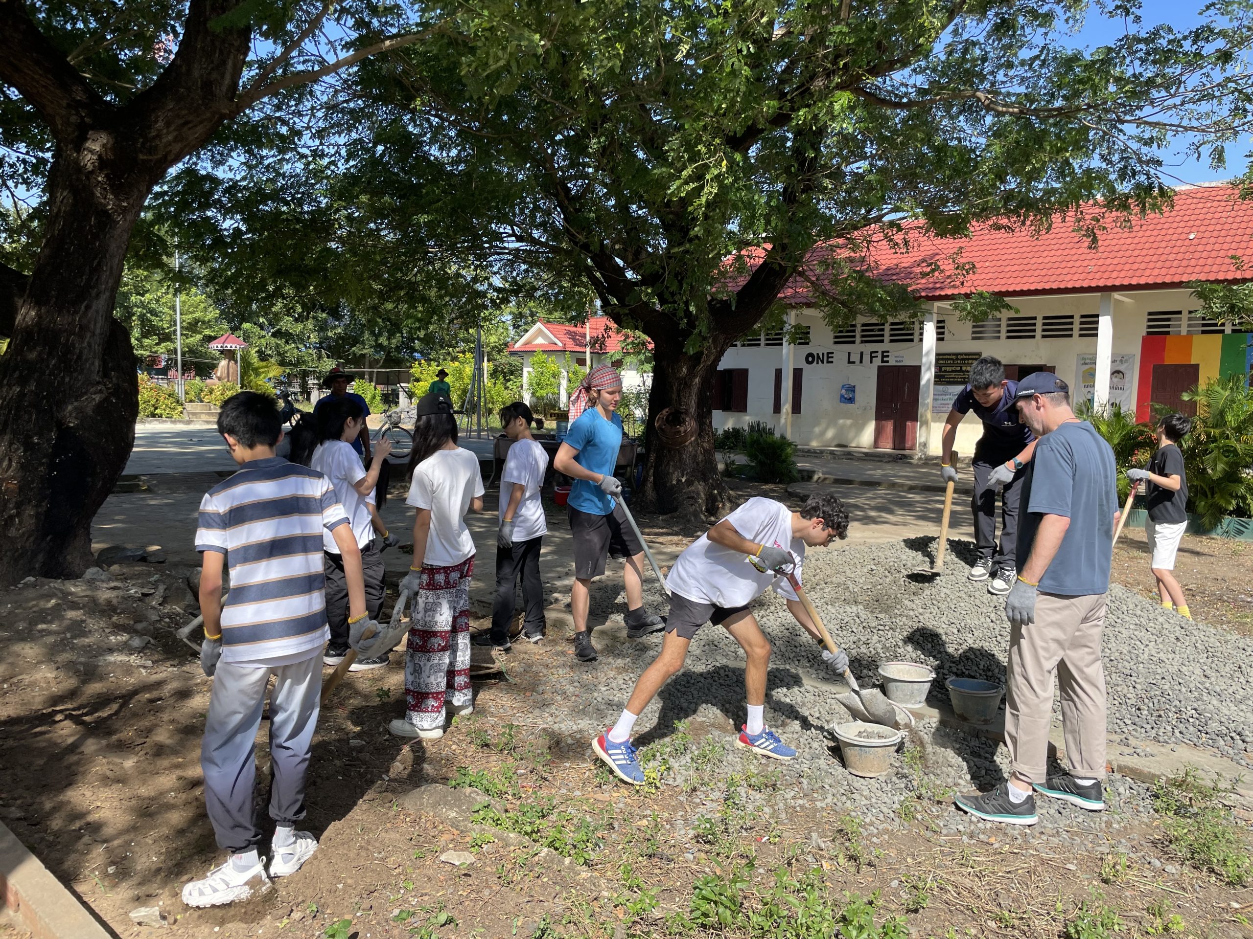 Students shovel gravel near a school building shaded by large trees