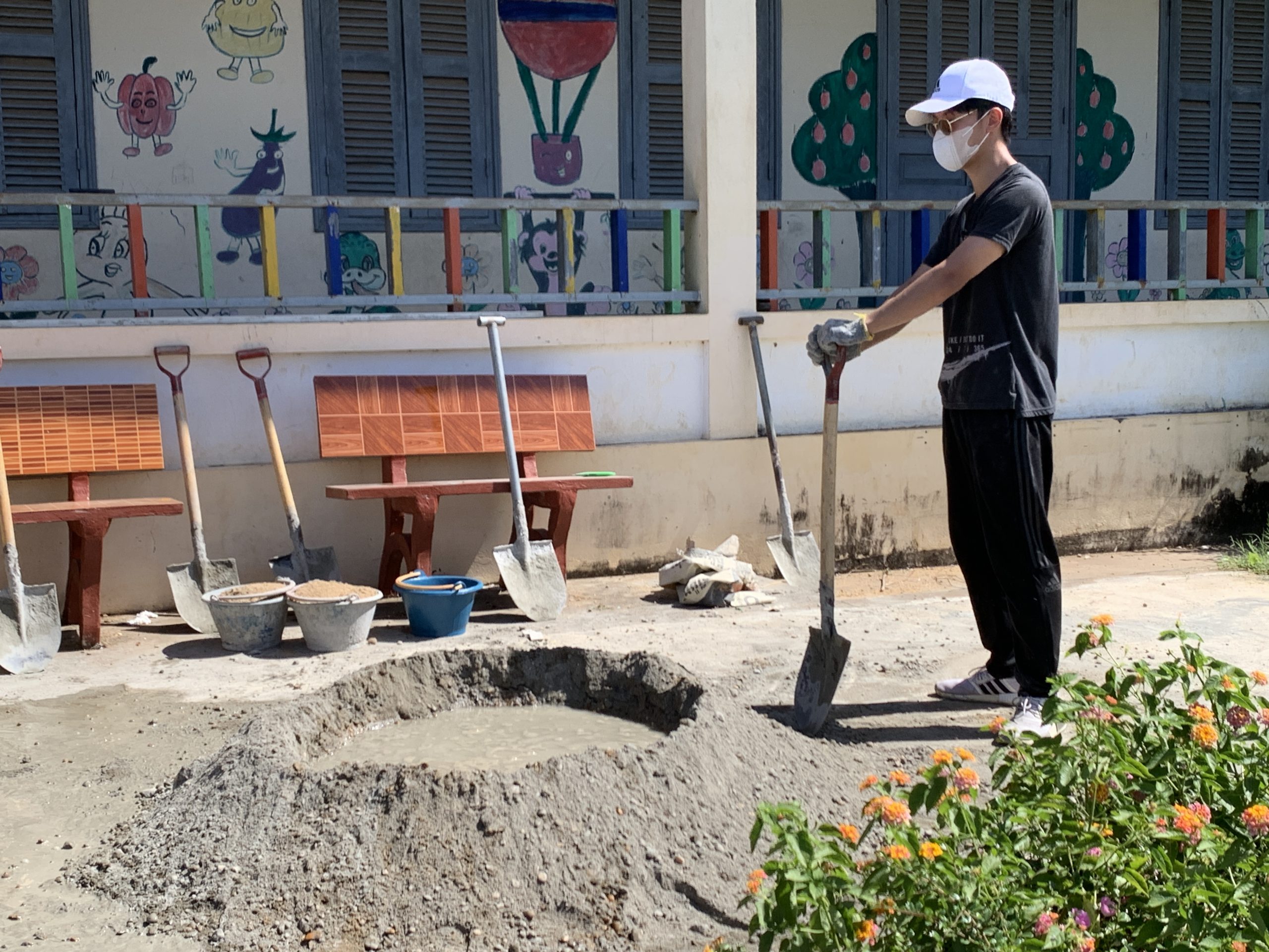 A student holds a shovel beside piles of sand and gravel at a school painted with colorful murals