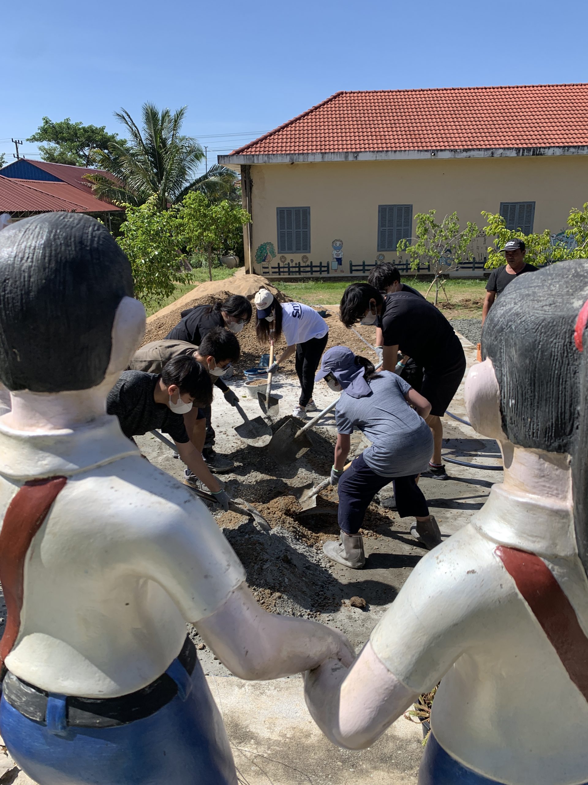 A school trip pauses for a community service project as students shovel and mix gravel, framed by two painted statues