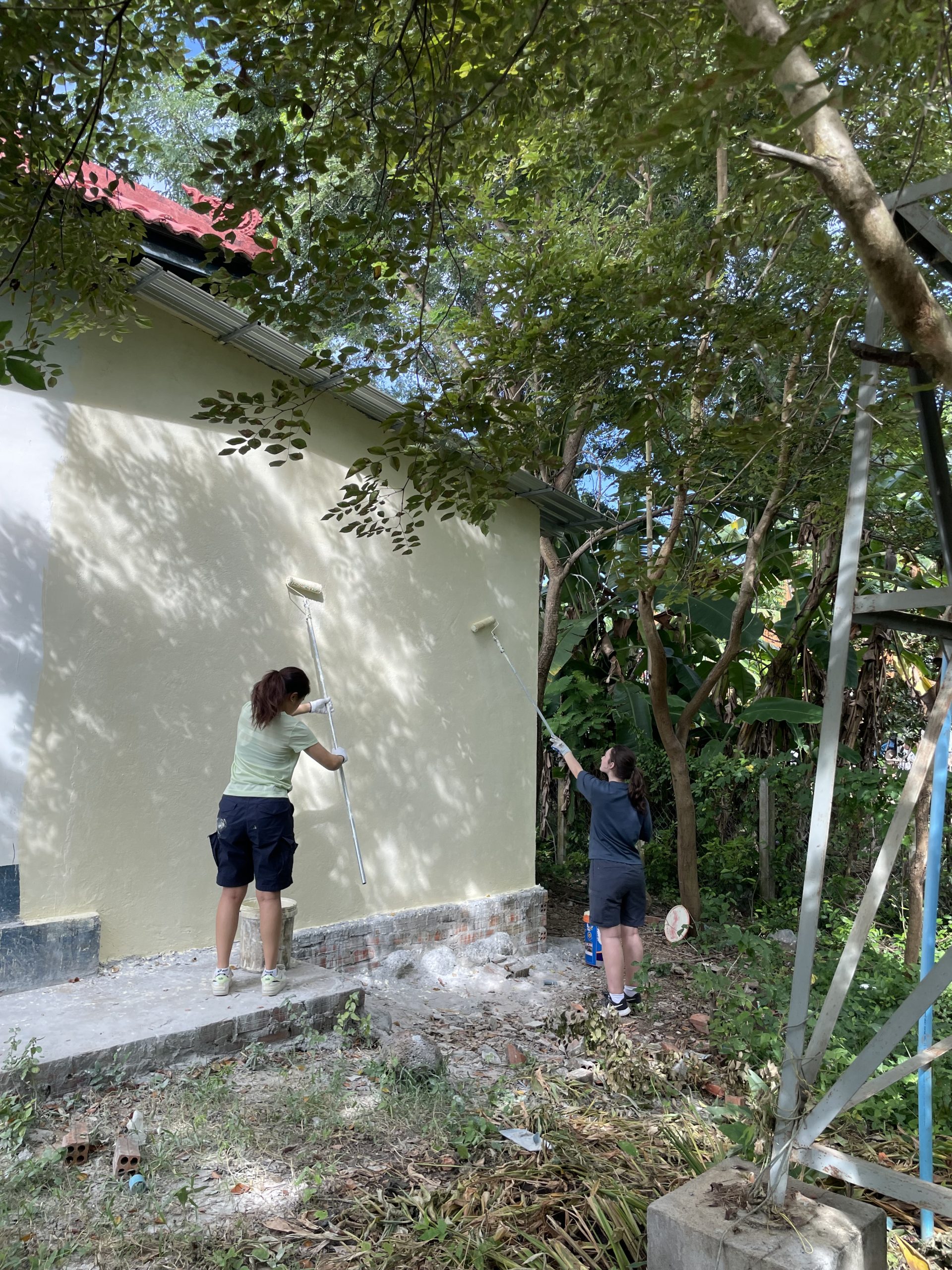 Students paint the exterior wall of a building with rollers under a tree canopy