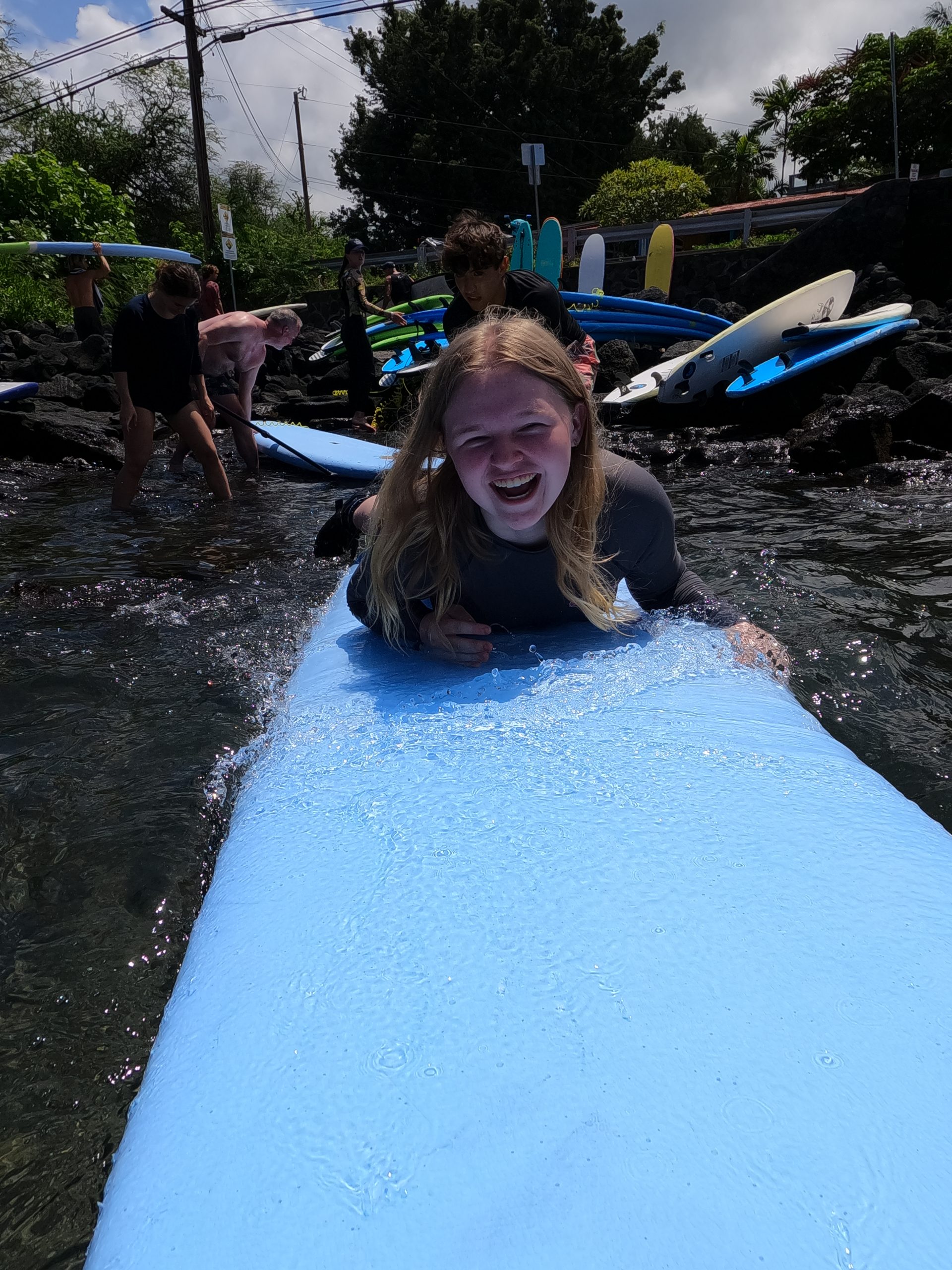 A student lies on a surfboard laughing, with others wading nearby among stacked boards