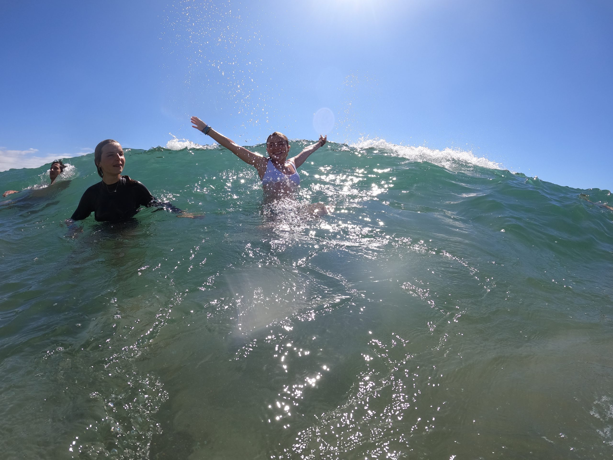 Students play in ocean waves, arms raised as a swell crests behind them