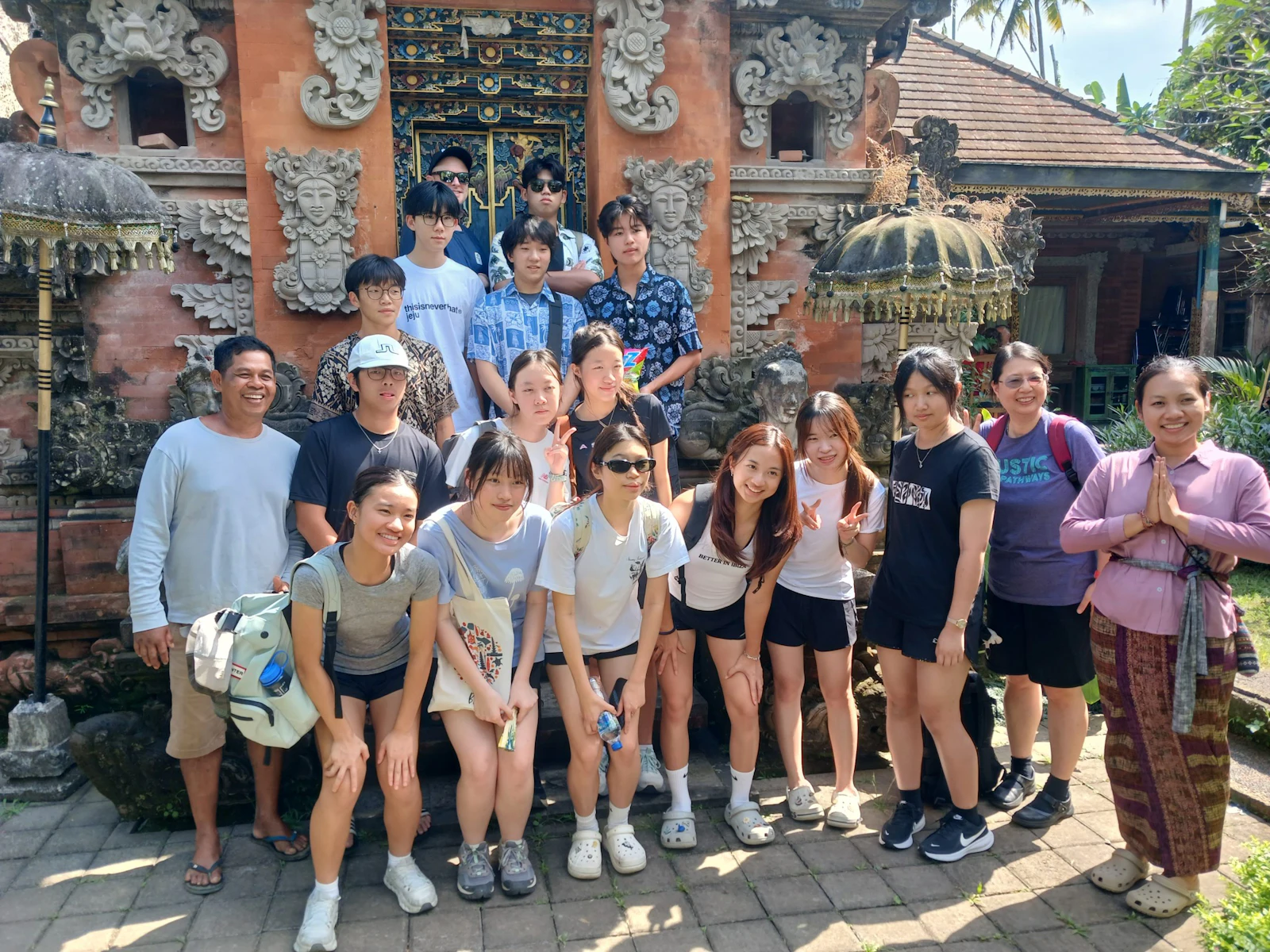 A group of teen travelers stands with three local hosts in front of a carved orange-brick temple gate in Bali, one host pressing her palms together
