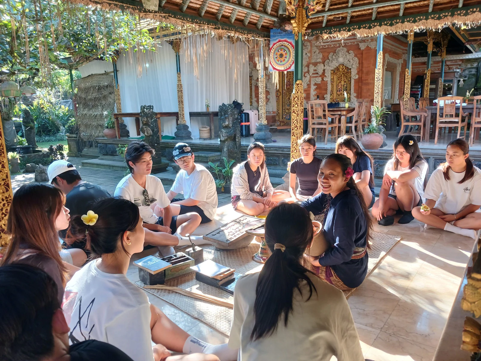 Teen travelers sit in a circle on a pavilion floor in Bali around a local teacher, with trees and a carved gate behind them