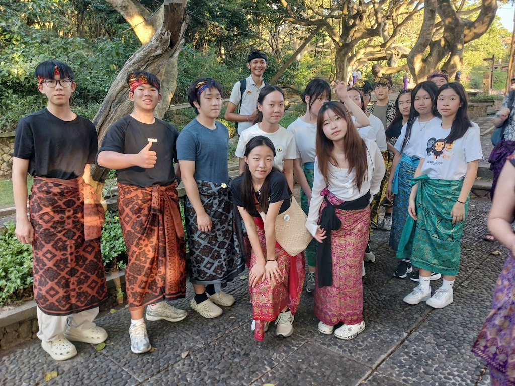 Teen travelers wearing traditional Balinese sarongs and headbands gather under trees at a temple in Bali.