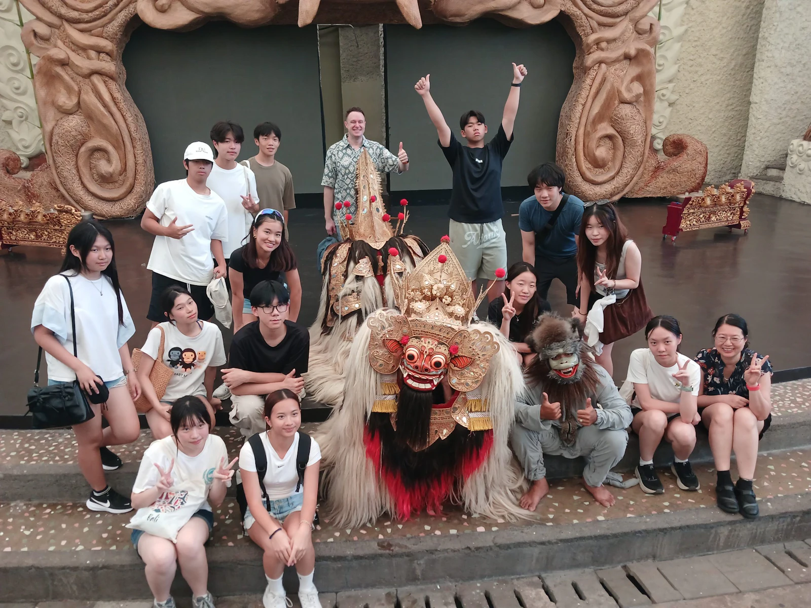 A group of teen travelers cheers with arms raised around Barong and Rangda performers on a stage in Bali