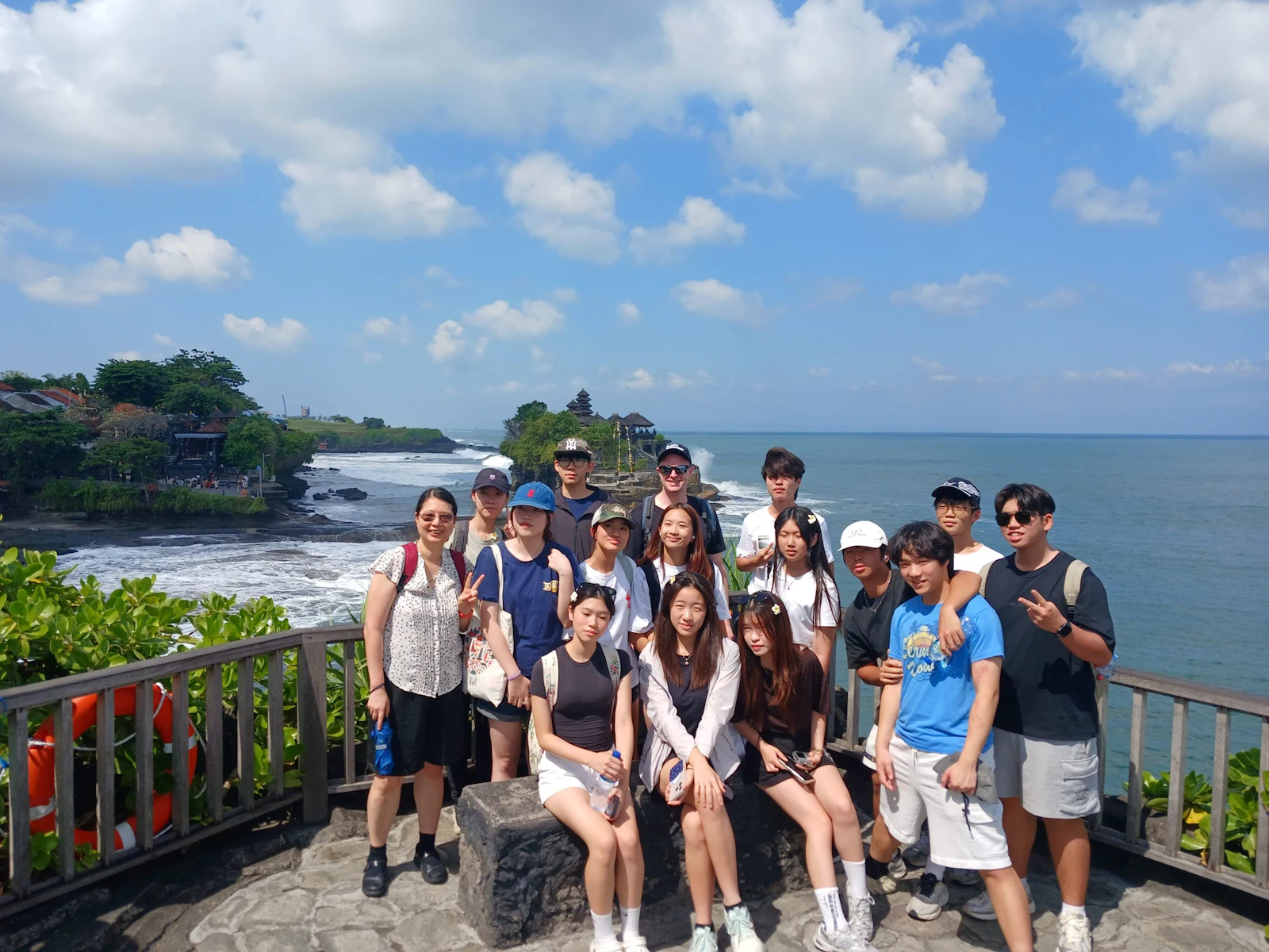 A group of teen travelers stand along a railing above the sea in Bali, a temple on a rocky headland behind them