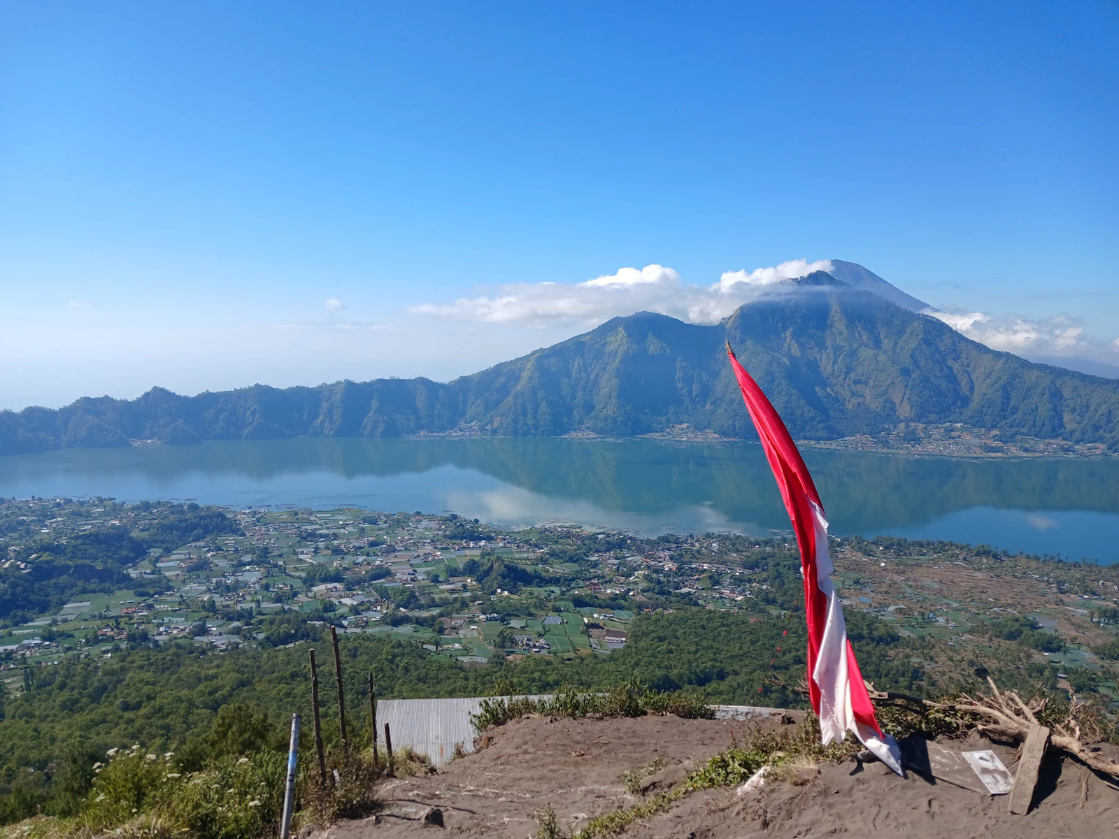 A volcano rises beyond a blue crater lake and a village in Bali, a red-and-white flag in the foreground