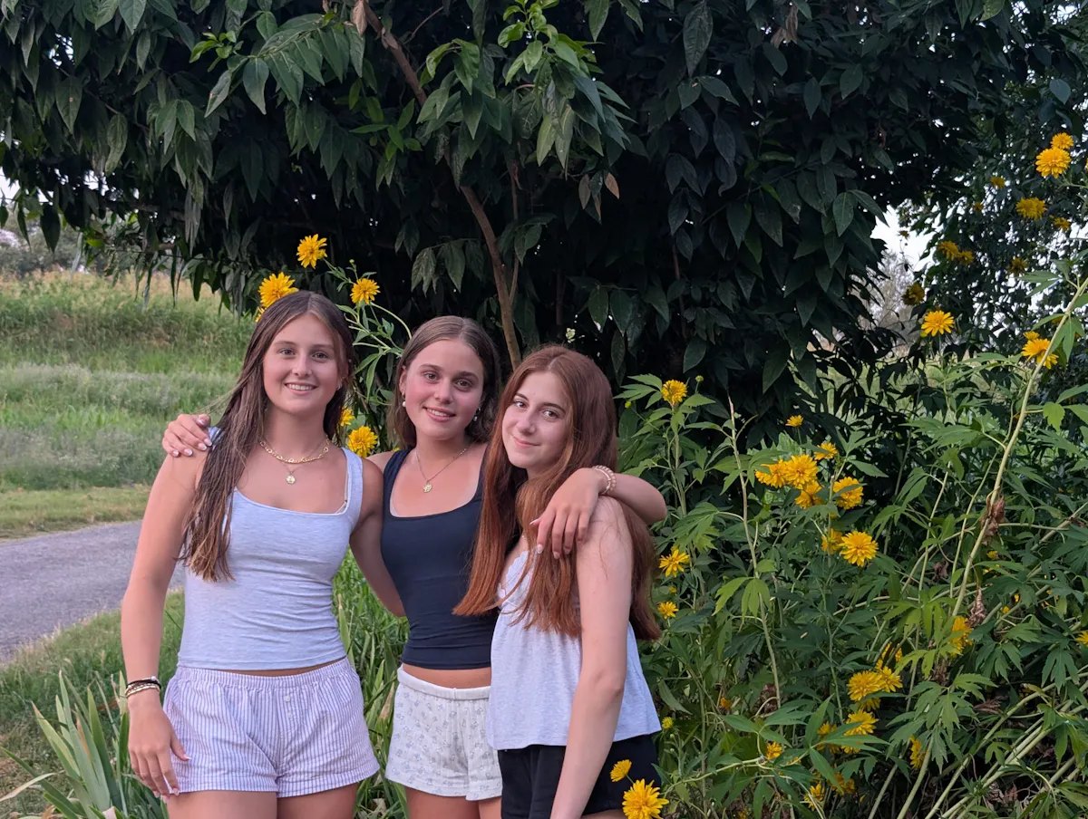 Three students standing arm in arm on a path beside a flowering shrub and a broad green tree