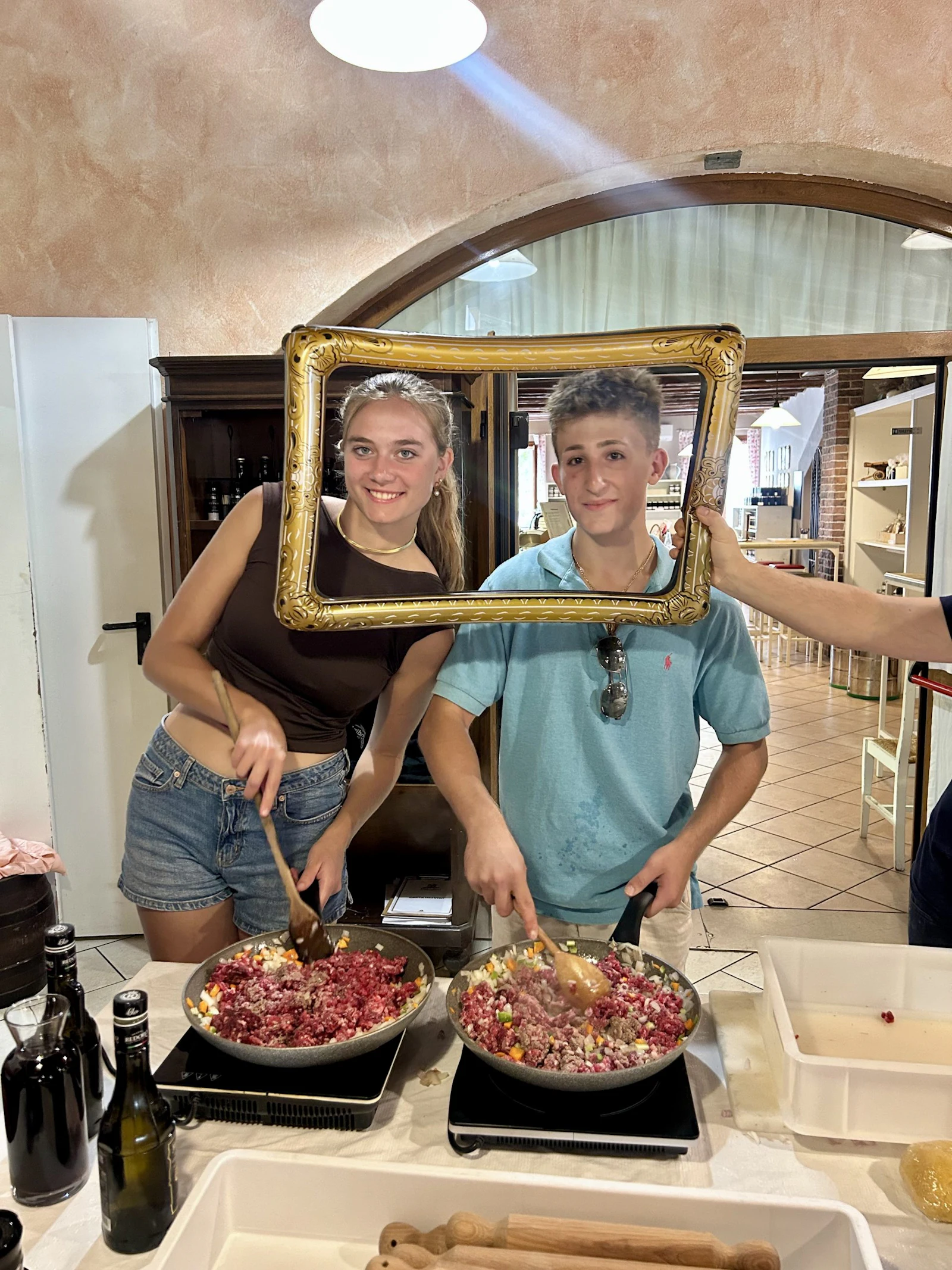 Two teen travelers hold up an empty gilded picture frame while stirring pans of sauce at a cooking class in Italy