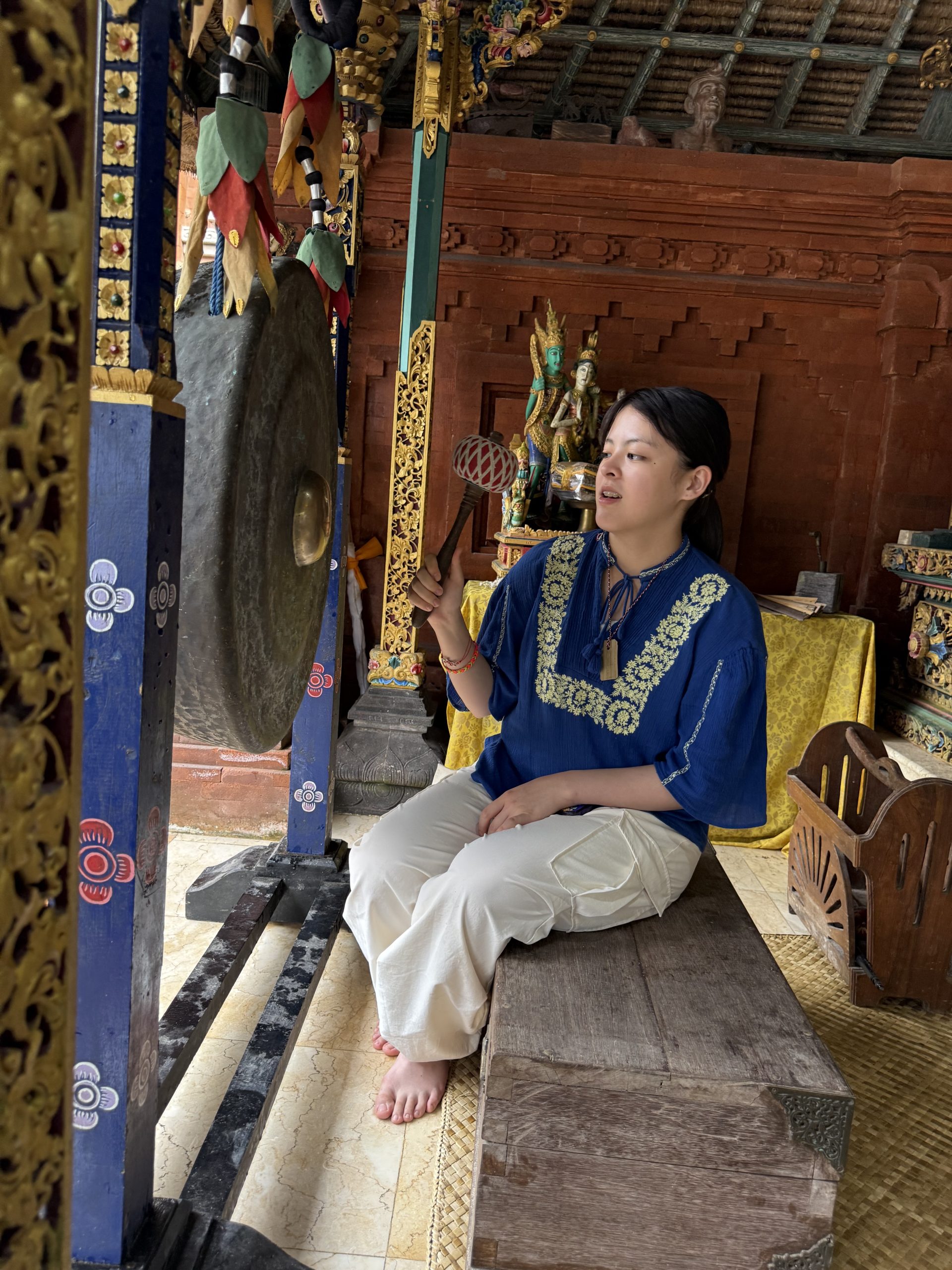 A student in traditional dress holds a mallet beside a large bronze gong inside an ornately decorated temple pavilion.