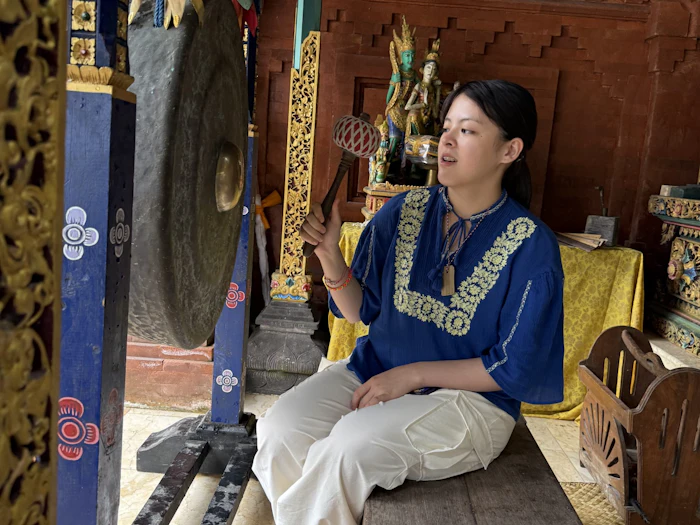 A student in traditional dress holds a mallet beside a large bronze gong inside an ornately decorated temple pavilion.