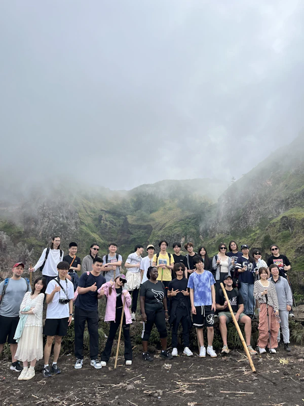 A large group of teen travelers stands together with walking sticks on a rocky ridge below a cloud-covered volcanic slope in Bali