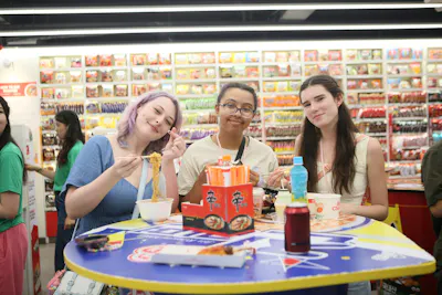 Three students share instant noodles at a table in a Seoul snack shop, shelves of packaged food behind them
